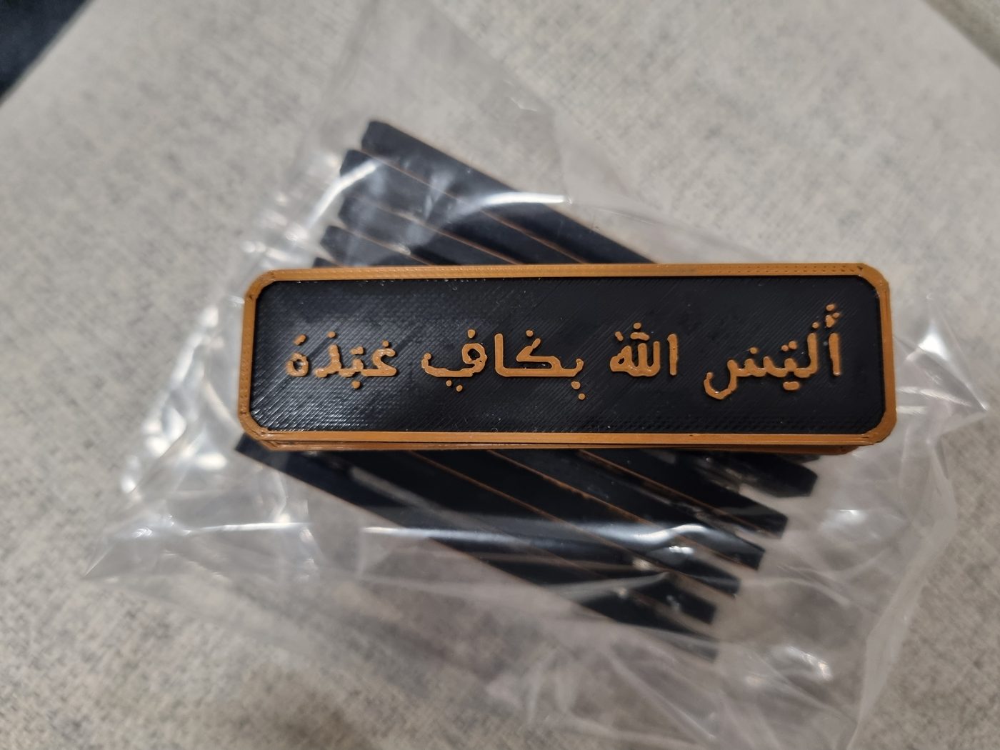 A rectangular black plaque with a gold border and raised gold Arabic calligraphy, on a clear bag of more black plaques.