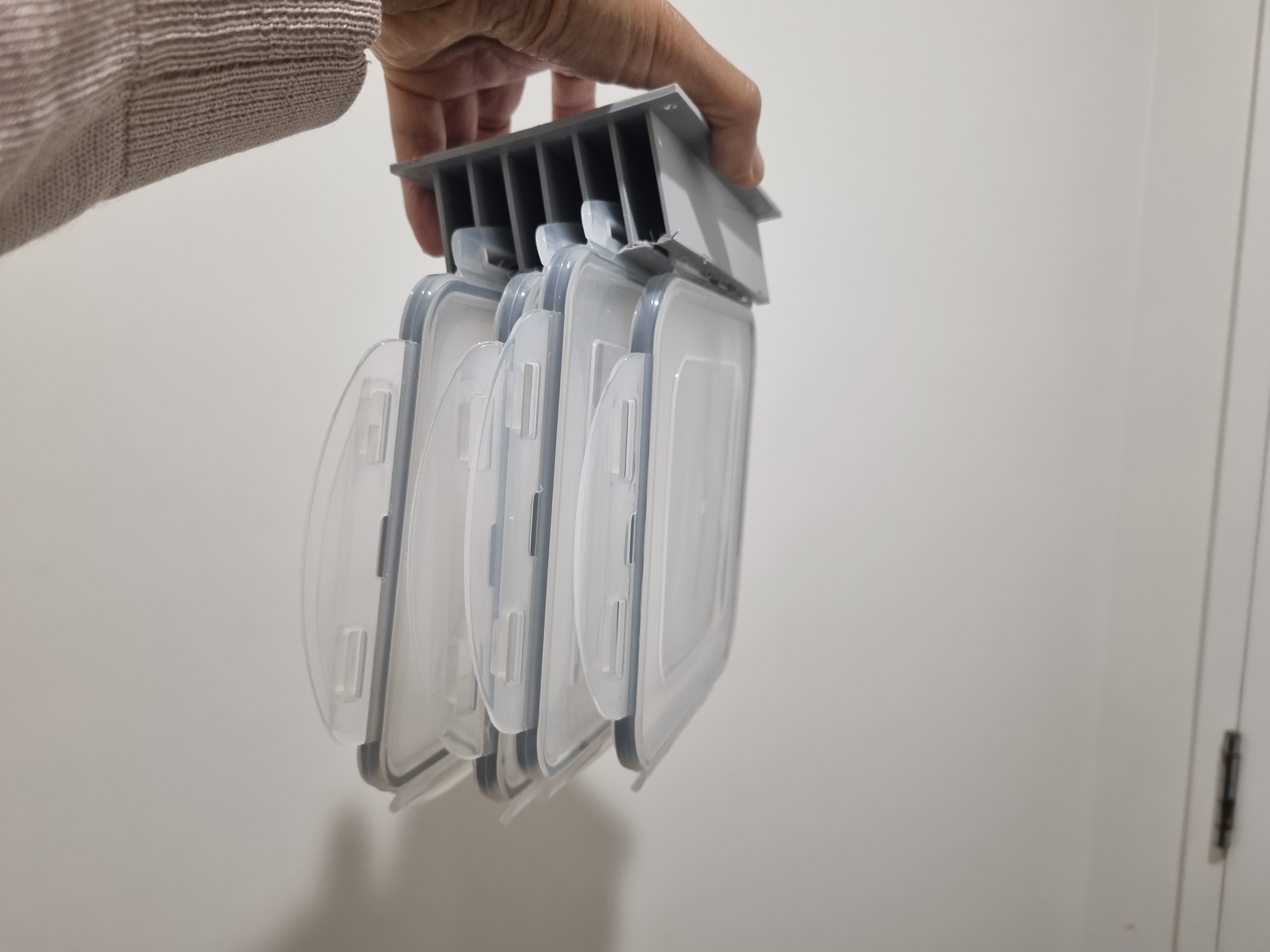 A grey 3D printed storage doodad, held up, with three clear kitchen container lids hanging from it
