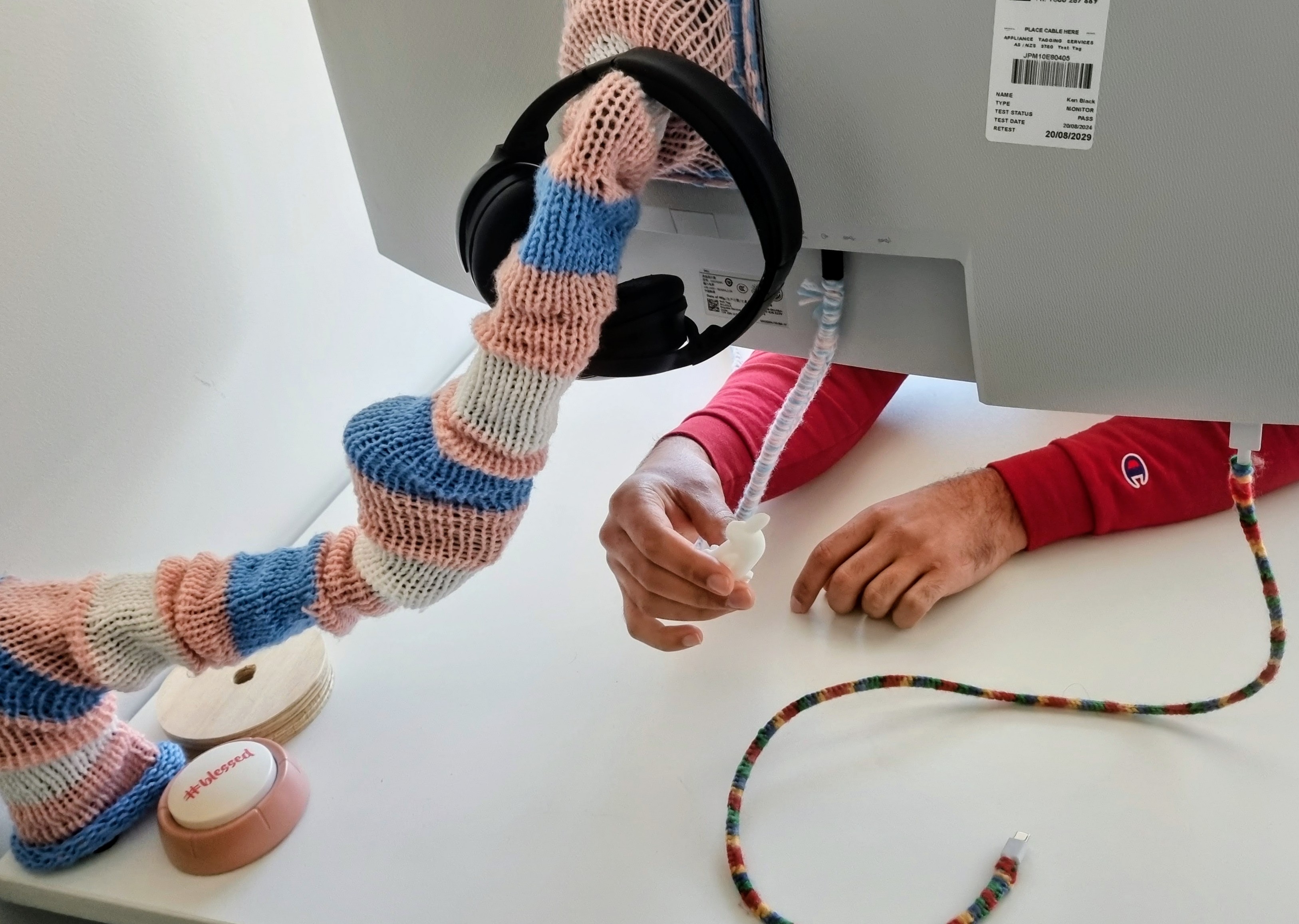 A pink, blue and white knitted sleeve on a monitor arm, with USB cables wrapped in coloured yarn