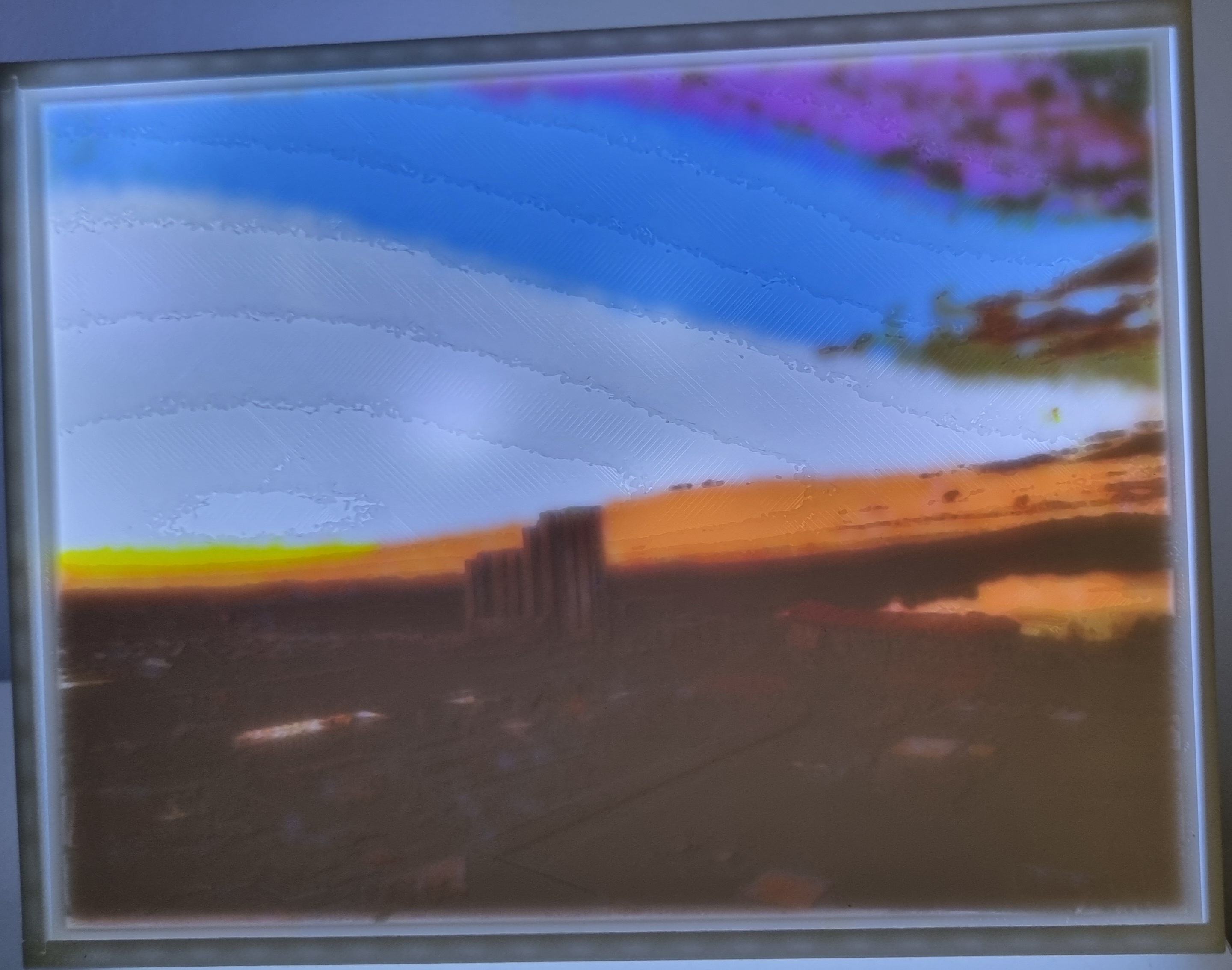 A lithophane of a city sunset, glowing warm orange in a white frame, with a lamp shining behind it