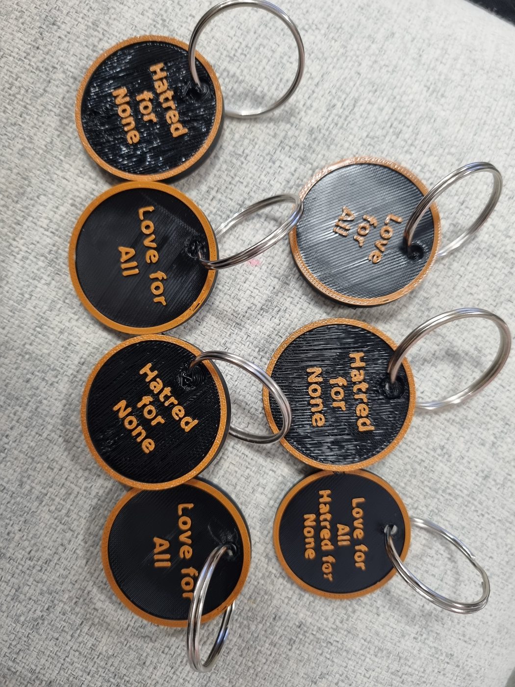 Round black keyrings with an orange-gold rim and raised gold lettering. Some read Love for All, some read Hatred for None, and one shows both.