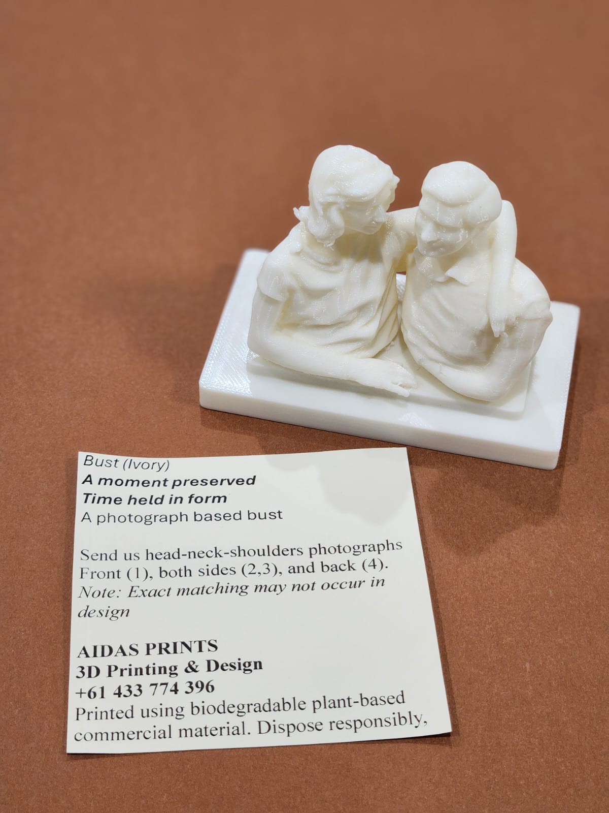 A small cream 3D printed bust of a smiling couple, next to an Aidas Prints card